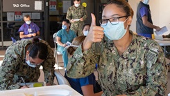 Several military personnel, wearing masks, filling out paperwork. One woman is giving the thumbs up sign