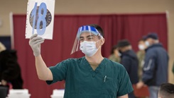 Military health personnel wearing a mask and a face shield holding up a sign that has the number eighteen on it 