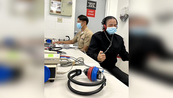 Image of Female service member in front holds a clicker while wearing a headset. In the background is the hearing test technician..